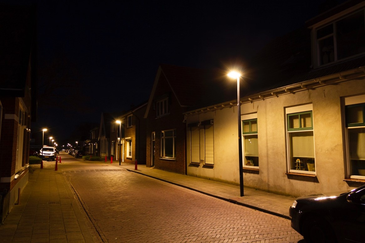 Straat in het donker met huizen aan beide kanten van de straat en straatverlichting. Zie bijschrift onder de afbeelding